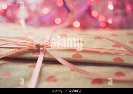 Geschenk in Papierverpackung mit einem Muster roter Herzen. Schleife mit Band und rosafarbene Lichter im Hintergrund Stockfoto