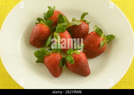 Frische rohe rote Erdbeeren mit Blättern in weißer Schüssel auf gelber Tischdecke Stockfoto