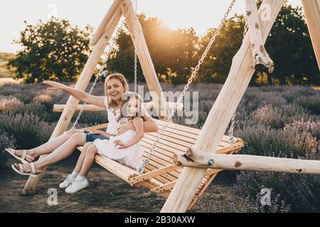 Schöne kaukasische Mutter und ihre kleine Tochter, die in einer Schaukel mit einem Lavendelfeld auf dem Hintergrund sitzen Stockfoto