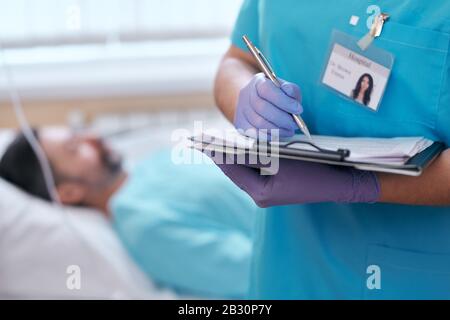 Nahaufnahme der nicht erkennbaren Krankenschwester in chirurgischen Handschuhen, die die Patientenkarte im Krankenhaus ausfüllen Stockfoto