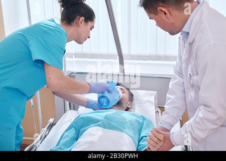 Die junge Krankenschwester, die den Patienten die Sauerstoffmaske der Beutelklappe aufsetzt, muss sein Puls überprüfen, während sie dem Menschen dringende Hilfe leistet Stockfoto