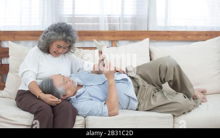 Älterer asiatischer Mann legt sich auf das Sofa, das Buch mit der Großmutter im Wohnzimmer zu Hause liest. Stockfoto