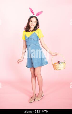 Studio voller Länge von asiatischer Schönheit fröhliche junge Frau, die bunte Ohren trägt und farbenfrohes Osterei in Holzkorb mit wunderschönem Lächeln und Colo hält Stockfoto