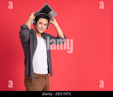 Junger Flusspferde gutaussehender asiatischer Typ mit kabellosen Ohrhörern auf dem Kopf, die Spiele auf einem Laptop-Computer spielen, während er isoliert auf Rot steht Stockfoto