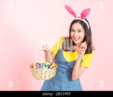 Studio in halber Länge von asiatischer Schönheit fröhliche junge Frau, die bunte Ohren trägt und mit einem schönen Lächeln buntes Osterei im Holzkorb hält Stockfoto