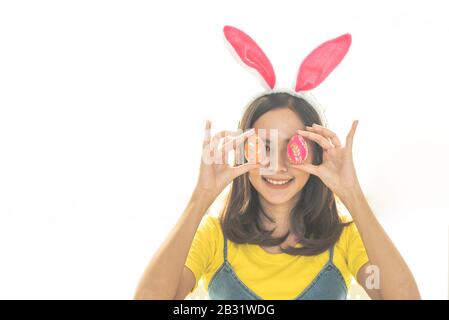 Studio von asiatischer Schönheit fröhliche junge Frau, die bunte Ohren trägt und ein buntes Osterei vor ihrem Auge hält Stockfoto