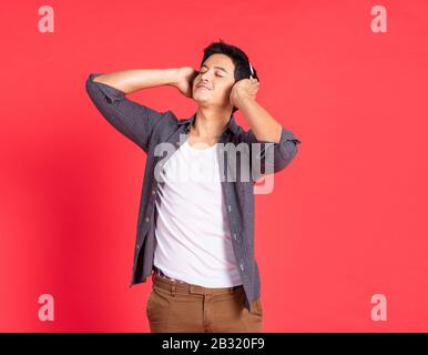 Bild eines fröhlichen jungen asiatischen Studentenkleides in legeren Hemdsingen und Musik hören mit Handy und schnurlosen Ohrhörern isoliert auf Rot Stockfoto