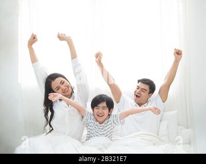 Fröhliche Familie wachen am Morgen auf, streckt die Hand hoch zur Luft, während sie im Schlafzimmer mit großem Fenster im Hintergrund auf dem Bett sitzt. Stockfoto