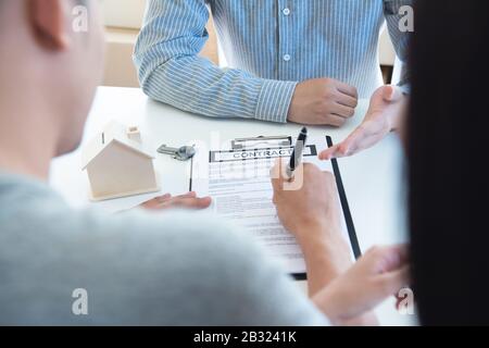 Glückliches junges asiatisches Paar und Realtoragent. Fröhlicher junger Mann, der zusammen mit seiner Frau einige Dokumente unterschreibt, während er am Schreibtisch sitzt. Neues Haus kaufen Stockfoto
