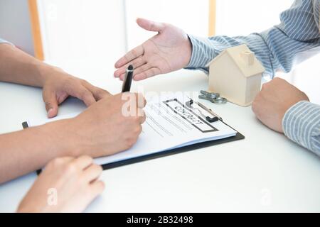 Glückliches junges asiatisches Paar und Realtoragent. Fröhlicher junger Mann, der zusammen mit seiner Frau einige Dokumente unterschreibt, während er am Schreibtisch sitzt. Stockfoto