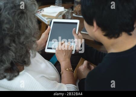 Rückansicht der älteren älteren älteren älteren älteren asiatischen Frau, die die Anwendung zur Gesundheitsüberwachung auf einem digitalen Tablet verwendet, während sie zusammen mit Grandson im Wohnzimmer versammelt ist Stockfoto
