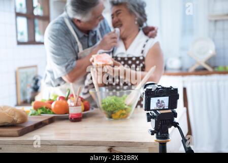 Paares älteres asiatisches älteres glückliches Leben in der Hausküche. Großvater wischt den Mund der Großmutter, nachdem er Brot mit Marmelade vlog vdo für Social Blogger gegessen hat. Stockfoto