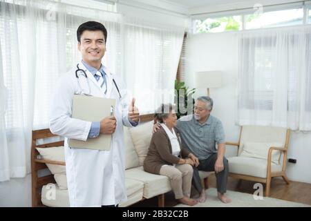 Junge gutaussehende kaukasische männliche Ärztin steht Hand in der Dokumentenakte Blick auf die Kamera mit Lächeln und zwei ältere alte ältere asiatische Paare sitzen auf dem Sofa Stockfoto