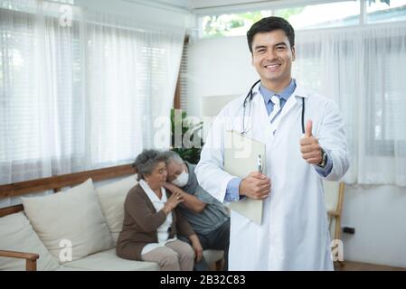 Junge gutaussehende kaukasische männliche Ärztin steht Hand in der Dokumentenakte Blick auf die Kamera mit Lächeln und zwei ältere alte ältere asiatische Paare sitzen auf dem Sofa Stockfoto