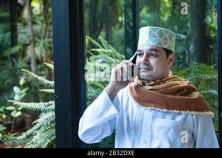 Mittelöstlicher Emirati-Mann in traditioneller Kleidung namens kandura auf dem Stuhl im Coffeeshop sitzen und mit seiner mobilen Kommunikation sprechen Stockfoto