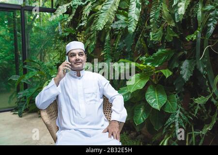 Mittelöstlicher Emirati-Mann in traditioneller Kleidung namens kandura auf dem Stuhl im Coffeeshop sitzen und mit seinem Mobile.Smart Communication Tech sprechen Stockfoto