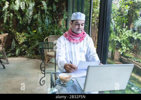 Junge gutaussehende arabische geschäftsleute im Café mit gekleideten Hijab sehen sich Papierakte mit Laptop auf dem Tisch an. Stockfoto
