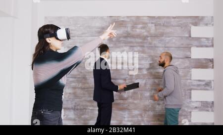 Designerin mit virtueller Reality-Brille in leerer Wohnung, während Immobilienmakler im Hintergrund mit dem Hausbesitzer spricht. Stockfoto