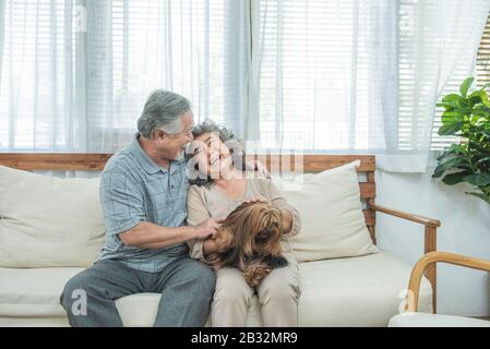 Glückliches älteres älteres asiatisches Paar sitzt auf dem Sofa zusammen mit Haustiertherapie in der Krankenpflege, pensionierter Mann und Frau, die Hund halten, während sie auf der Couch sitzen Stockfoto