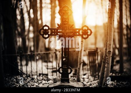 Auf einem steinernen Grabstein in der Mitte des Friedhofs steht ein schwarzes altes gemustertes Kreuz, umgeben von einem dunklen Wald, durch dessen Traufen die br Stockfoto