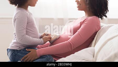 Kleine Tochter, Die Kleine Baby-Schuhe Auf Den Bauch Der Schwangeren Afro Mutter Legt Stockfoto