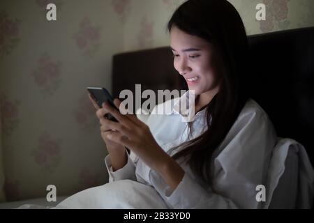 Asiatische Frau, die nachts ein Handy am Bett benutzt.Sie lächelt und genießt zu Hause. Stockfoto