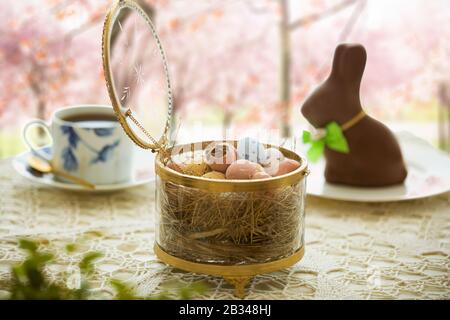 Schöner, kleiner Tisch mit Osterdekorationen auf der Außenterrasse. Kleiner Schokoladenbrötchen mit Schleife, Schokoladeneiern, Tasse Kaffee, Kristallschale. Stockfoto