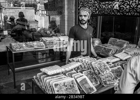 Aqaba, JORDANIEN - 31. JANUAR 2020: Junger Mann mit Bart und arabischem Kopftuch verkauft im sonnigen Wintertag draußen auf der Straße Gebäck, öffentlicher Stadtmarkt. Schwarz-Weiß-Bild Stockfoto