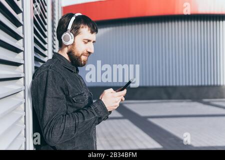 Junger bärtiger Mann, der Musik in kabellosen Kopfhörern in der städtischen Umgebung hört und sein Handy in den Händen hält Stockfoto