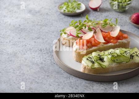 Drei Sandwiches auf Ciabatta Toast mit frischem Gemüse, Radieschen, Tomaten, Gurken und Mikrogrüns auf grauem Hintergrund. Nahaufnahme. Kopierbereich. Stockfoto
