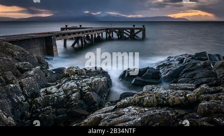 Der alte Steg in Portencross in Schottland und die Winterhügel auf der Insel Arran, während die Sonne untergeht und einen dramatischen Himmel über dem Sonnenuntergang erzeugt. Stockfoto