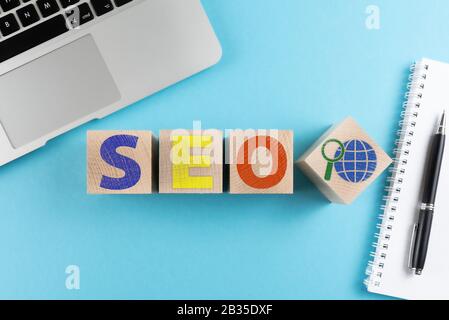 Draufsicht über die Buchstaben SEO und Internetsuchsymbol auf Holzwürfeln auf blauem Hintergrund mit Laptop und Spiralnotizbuch Stockfoto