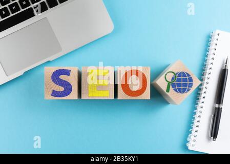 Draufsicht über die Buchstaben SEO und Internetsuchsymbol auf Holzwürfeln auf blauem Hintergrund mit Laptop und Spiralnotizbuch Stockfoto