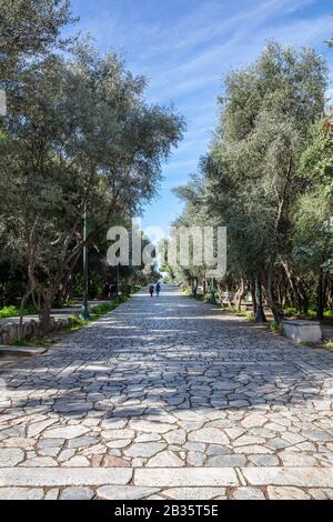 Gepflasterter Fußweg aus altem Stein am Filopappou-Hügel in Athen, Griechenland. Die Menschen gehen an einem sonnigen Tag einen Kopfsteinpflaster unter der alten Akropolis. Bäume um, ve Stockfoto