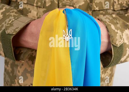 Ukrainischer Mann-Krieger, der mit einer Militär-Pixel-Uniform bekleidet ist, hält die gelb-blaue Flagge des Staates Ukraine und an der Kette ein kleines Wappen von Stockfoto