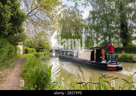 Yelvertoft, Northamptonshire, Großbritannien, Mai 2019: Ein Mann in einer wasserdichten Jacke lenkt ein schmales Boot in Richtung einer Brücke am Canal Grand Union. Stockfoto