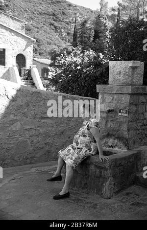 Die alte Frau ruht auf einem Brunnen, der Rue de la Fontaine du Portal, Saint-Guilhem-le-Désert; Hérault, Occitanie, Frankreich. Schwarzweiß-Version Stockfoto