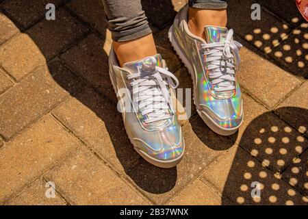 Zwei weibliche Füße mit bunt schillernden Turnschuhen bei strahlendem Sonnenschein, die Schatten auf Ziegelsteinfertigern erzeugen. Stockfoto