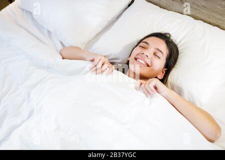 Fröhliche junge Frau im Bett mit weißen, sauberen Bettlaken, die lächeln und sich mit Decke bedecken. Bequeme Matratze Stockfoto