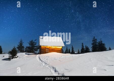 Fantastische Winterlandschaft mit Holz- Haus in Snowy Mountains. Rauch kommt aus dem Schornstein der verschneite Hütte. Weihnachten und Winter Ferien Konzept Stockfoto