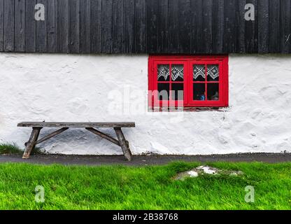 Weiße Hauswand mit rotem Fenster auf altem färöischem Haus. Holzbank auf grünem Gras. Faröer Inseln, Dänemark Stockfoto