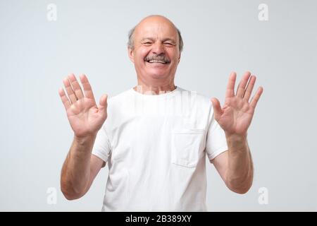 Aufgeregter kahler Mann zeigt zehn Finger und lacht freudig isoliert auf weißem Hintergrund, legt die Hände hoch, als er bittet, ihn nicht mehr zum Lachen zu bringen. Studio gedreht Stockfoto