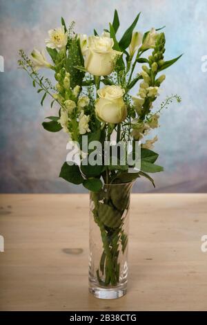 Weiße Rosen und verschiedene weiße Blumen in einer Anordnung in einer Glasvase auf einem Kieferntisch. Stockfoto