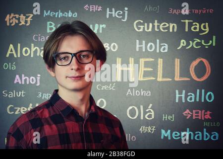 Positiver Teenager, der rotes Hemd und Brille über grauem Wandhintergrund trägt und das Wort Hallo in verschiedenen Sprachen und Farben geschrieben. Lernende OP Stockfoto