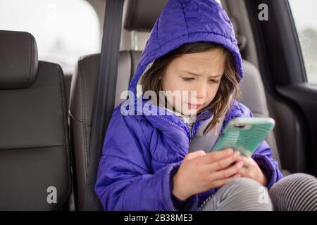 Ein kleines Mädchen spielt mit Telefon auf der Rückbank eines Autos auf einer Autofahrt Stockfoto