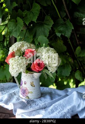 Sommer-Blumenstrauß aus Rosen und Hydrangeen in einer Vintage-Kanne auf dem Hintergrund eines grünen Gartens an einem sonnigen Sommertag. Nahaufnahme. Stockfoto