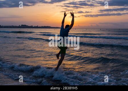 Silhouette des glücklichen fröhlichen Mann springen und Spaß am Strand gegen Sonnenuntergang. Das Konzept von Freiheit und Entspannung. Stockfoto