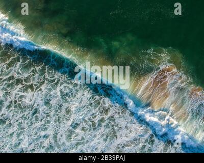Drone Blick auf schöne türkisblauen Wellen brechen auf sandigen Küste. Luftaufnahme von goldenem Strand, tiefblauem Meerwasser und schäumenden Wellen Stockfoto