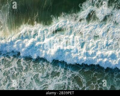 Drone Blick auf schöne türkisblauen Wellen brechen auf sandigen Küste. Luftaufnahme von goldenem Strand, tiefblauem Meerwasser und schäumenden Wellen Stockfoto