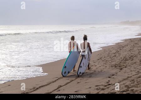 Zwei Surfer-Paare gehen mit Longboards in der Hand am Meeresstrand. Stockfoto